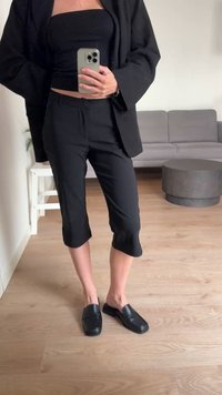 Calças pretas variadas com uma textura suave, combinadas com um top preto ajustado e sapatos pretos de sapatilha. O conjunto apresenta elementos de design minimalistas.