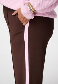 Bruine broek met een roze zijstreep, met een gladde textuur en een zijzak. Bovenaan is een roze, gebreide trui zichtbaar.