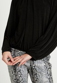 Czarna, teksturowana bluzka z długim rękawem i marszczonymi mankietami, zestawiona z dopasowanymi spodniami w wężowy wzór w odcieniach szarości i bieli. Ręce złożone na biodrach.