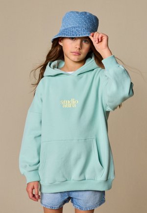 Jeune fille portant un sweat à capuche "studio aura" bleu clair, un short en jean et un bob en denim bleu, tenant le bord du chapeau d'une main.