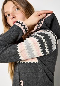 Sudadera de punto gris con patrón en zigzag en rosa, negro y gris en las mangas. Cuenta con capucha con cordón y un bolsillo frontal tipo canguro.