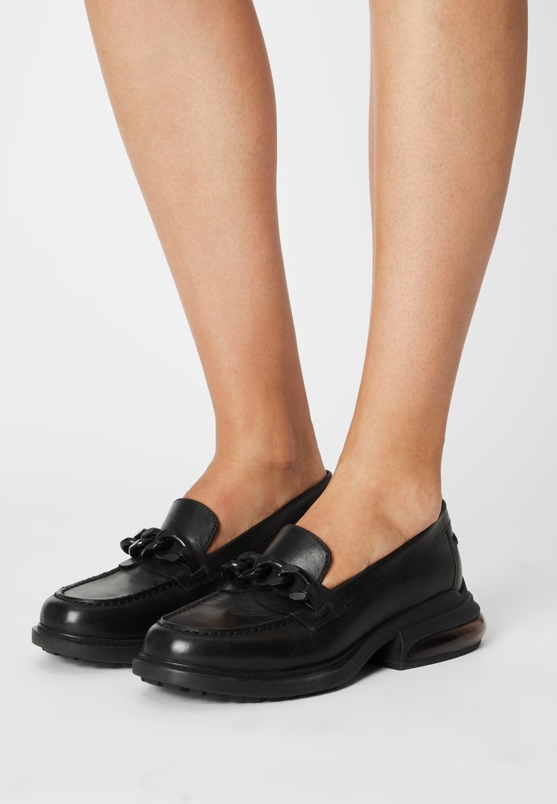 Mocassins en cuir noir avec détail de chaîne épaisse portés pieds nus, présentant des semelles épaisses et des bouts arrondis.