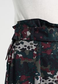 Jupe à motif camouflage dans des tons de vert, bordeaux et gris. Présente une taille large avec une ceinture en tissu assorti et des plis.