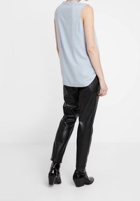Persona che indossa un top senza maniche di colore azzurro chiaro, pantaloni neri in pelle sintetica e stivali con tacco neri, in piedi contro uno sfondo bianco.