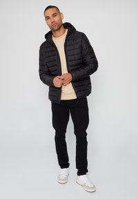 Zwarte pufferjack met een gewatteerde textuur en een aangehechte capuchon, gedragen over een beige top. Het model combineert het met zwarte broeken en witte sneakers.