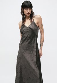 Sieviete ar tumšiem matiem un tetovējumiem, valkājot kroplētu melnu satīna slip kleitu ar plānām lenciņām uz vienmērīga gaiša fona.