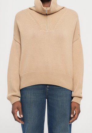 Personne portant un pull en maille beige avec un col montant et une demi-fermeture éclair, associé à un jean en denim bleu foncé, montré des épaules aux hanches.