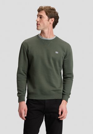 Uomo che indossa una felpa verde oliva con il logo Lee sul petto, guarda a sinistra, in piedi davanti a uno sfondo semplice grigio chiaro.