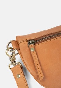 Tan leren handtas met een afgeronde vorm, voorzien van een ritssluiting, metalen hardware en een afneembare schouderriem met klinknagels en een musketonhaak.