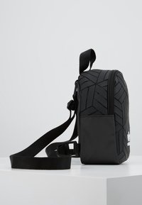 Svart axelremsväska med ett geometriskt texturerat mönster, stadigt material, dragkedjestängning och justerbar rem. Har märkning på framsidan.