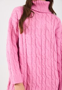Roza džemper s kabelskim pletenjem i visokim turtleneck-om, s rebrastim manžetama i teksturiranim uzorkom. Materijal izgleda mekano i debelo.