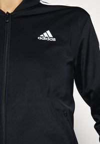 Czarna kurtka na zamek marki Adidas z białym logo na piersi i białymi paskami na ramieniu, noszona przez osobę na jednolitym tle.