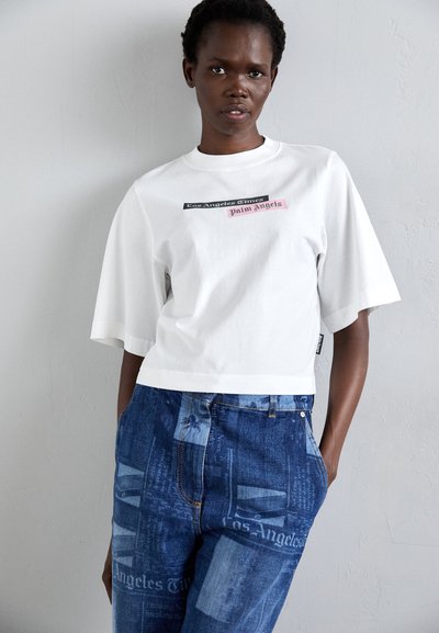 White cropped T-shirt with short sleeves featuring graphic text. Paired with blue jeans displaying a textured newspaper print pattern.