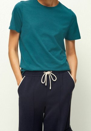 Personne portant un t-shirt à manches courtes bleu canard et un pantalon bleu marine avec un cordon blanc, mains dans les poches, sur un fond clair.