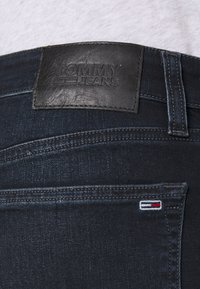 Jeans de mezclilla oscura con una etiqueta de cuero negro grabada con "TOMMY JEANS". Presenta costuras visibles y un pequeño logo bordado en rojo, blanco y azul.