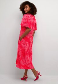 Dlouhá růžová šaty s paisley vzorem, rozšířenými rukávy, bočními kapsami a uvolněným střihem, doplněné o růžové vysoké podpatky. Hladké pozadí.