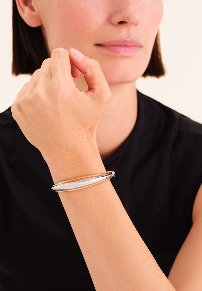 Bracelet manchette en métal argenté et doré avec une forme élégante et allongée et une texture lisse, porté au poignet contre une chemise noire.