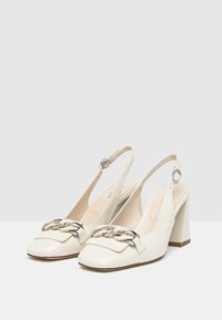 Bia�łe lakierowane szpilki typu slingback charakteryzują się kwadratowym noskiem, blokowym obcasem oraz dekoracyjnym łańcuszkiem na przodzie.
