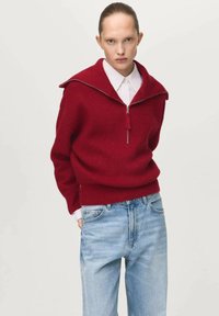 Roter Strickpullover mit weitem Kragen, halbem Reißverschluss vorne und gerippter Struktur, kombiniert mit hellblauen Denim-Jeans.