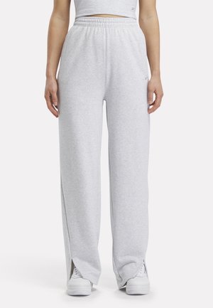 Frau trägt hellgraue Sweatpants mit weitem Bein und weiße Turnschuhe, auf Taille beschnitten, vor einfachem hellem Hintergrund.