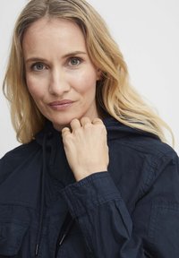 Jūras zilas krāsas jaka ar kapuci un kabatām, ar vieglu, saburzītu tekstūru. Modelis valkā vairākus zelta gredzenus uz pirkstiem.