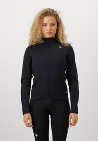 Žena s kučeravými vlasmi, oblečená v čiernej bundě na zips a čiernych cyklistických legínach, stojí proti jednému svetlému pozadiu.