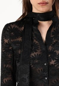 Black sheer blouse with floral embroidery, features a button-up front and a matching scarf tied around the neck.