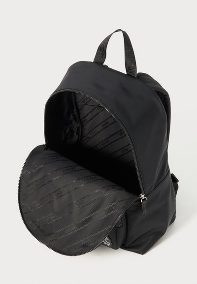 Rucsac negru cu un material mat, neted și o formă rotunjită. Dispune de un sistem de închidere cu două fermoare și un interior cu o căptușeală cu un model care afișează "Versace."