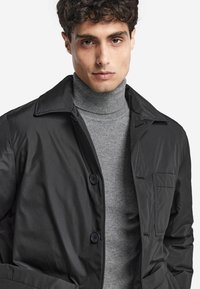 Czarna wodoodporna kurtka z kołnierzem, dwoma guzikami z przodu i kieszenią na piersi, noszona na jasnoszarym golfie. Gładka faktura.