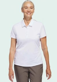 Λευκό πόλο μπλουζάκι με γιακά και πέντε κουμπιά, φτιαγμένο από απαλό ύφασμα. Φορεμένο με καφέ παντελόνι. Το μοντέλο έχει κοντά, ανοιχτόχρωμα μαλλιά.