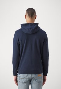 Felpa blu navy con cappuccio regolabile, texture morbida, maniche lunghe e polsini a coste. Abbinata a jeans chiari strappati.