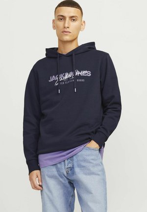 Navy hoodie made of soft fabric, featuring a white and purple logo print. Worn over a purple layer and paired with light blue jeans.