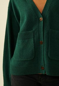 Grön stickad cardigan med v-ringad hals, träknapp, två framfickor och ribbade muddar. Bärs över en brun ribbad topp.