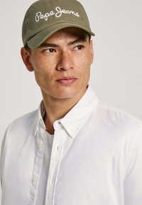 Olivno zelena kapa z belim vezenjem "Pepe Jeans", nosi se z belo srajco s gumbi čez sivo majico, ki ima gladko teksturo.