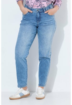 Persoon draagt lichtblauwe spijkerbroek, witte sneakers met blauwe strepen en een roze shirt met patroon en lange mouwen, handen in de zakken.