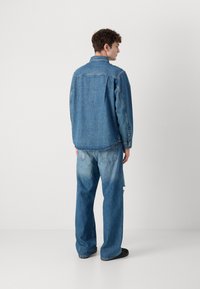 Camisa de mezclilla azul clara con cuello, cierre de botones en la parte delantera y mangas largas. Combinada con jeans de mezclilla azul desgastados y de corte suelto.