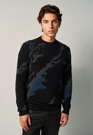Junger Mann mit kurzen dunklen Haaren, trägt einen schwarzen Pullover mit grauen und blauen abstrakten Mustern und schwarze Hose, steht vor einem einfarbigen Hintergrund.