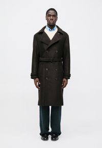 Abrigo marrón de lana con doble botonadura, solapas anchas, cinturón a juego y dos bolsillos laterales, superpuesto sobre un suéter claro y una camisa azul.