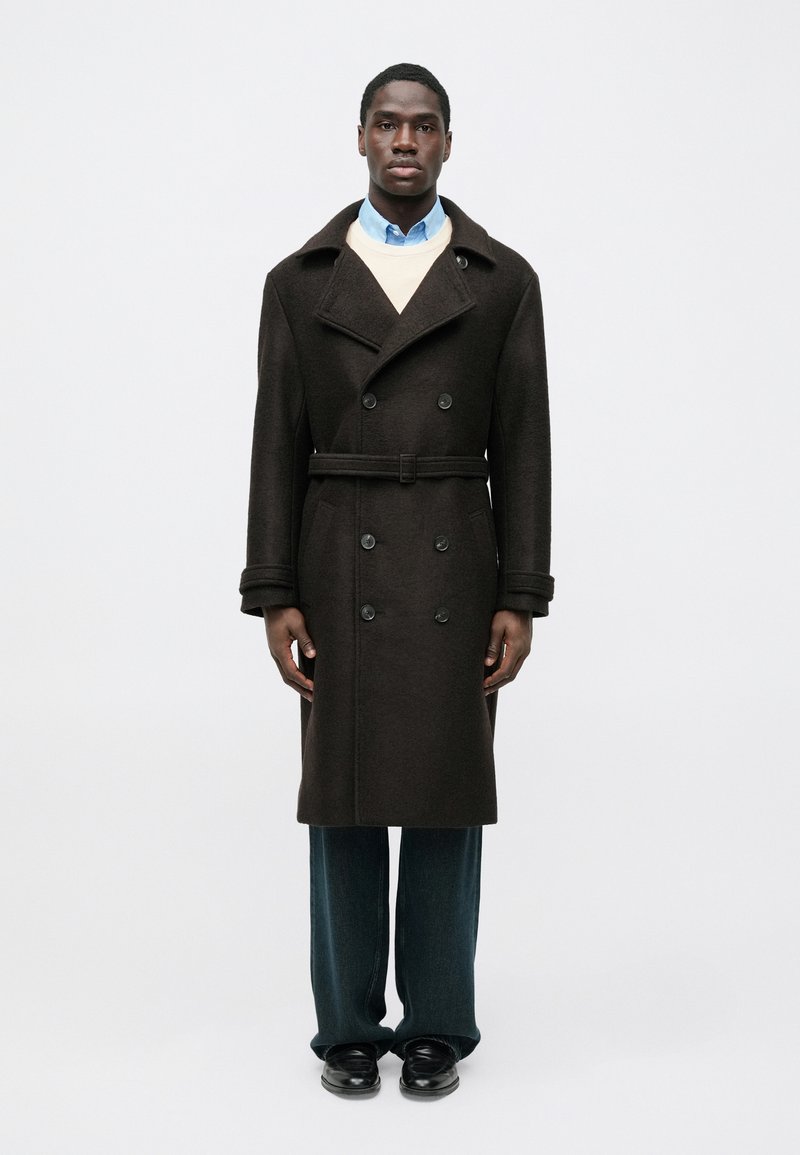 Abrigo marrón de lana con doble botonadura, solapas anchas, cinturón a juego y dos bolsillos laterales, superpuesto sobre un suéter claro y una camisa azul.