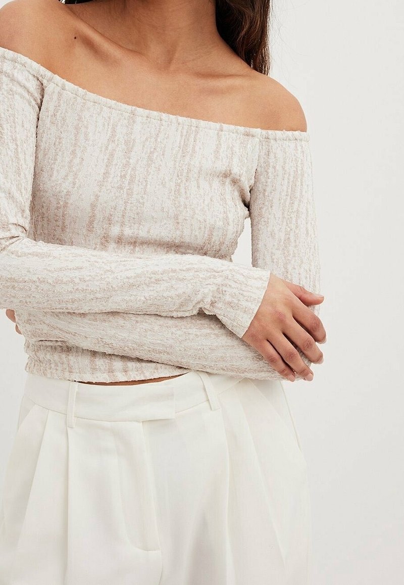 Kvinna som bär en texturerad off-shoulder långärmad beige topp och högmidjade veckade vita byxor med händerna korsade vid midjan.