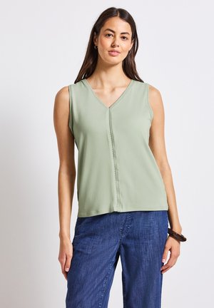 Top verde senza maniche con scollatura a V, caratterizzato da un dettaglio decorativo lungo il davanti e una finitura in tessuto liscia. Abbinato a pantaloni blu scuro.