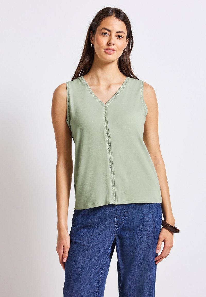 Top verde senza maniche con scollatura a V, caratterizzato da un dettaglio decorativo lungo il davanti e una finitura in tessuto liscia. Abbinato a pantaloni blu scuro.