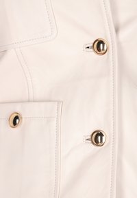 Hvid læderjakke med to guld-tonede knapper, sidelommer og synlige syninger. Glat tekstur med minimalistiske designdetaljer.