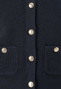 Marine blauwe gebreide cardi met ribstructuur, zes gouden knopen en twee voorkappen met knoopaccenten.