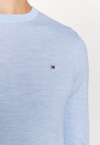 Un pull bleu clair à col ras du cou affichant un petit logo rectangulaire brodé rouge, blanc et marine sur la poitrine.