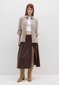 Gonna lunga a quadri marrone con uno spacco alto fino alla coscia, abbinata a un top bianco corto e una camicia a bottoni a quadri marrone, indossata con stivali beige con tacco.