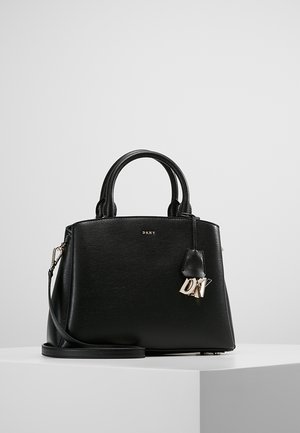Sac à main structuré noir DKNY avec poignées supérieures, bandoulière amovible et pendentif logo en métal DKNY sur une surface blanche.