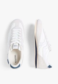 Zapatillas blancas que combinan materiales de cuero y gamuza, con punta redonda, diseño de cordones y un acento en azul marino en el talón.
