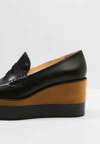 Mocassin à plateforme compensée en cuir noir avec une semelle marron et une bride décorative à l'avant.