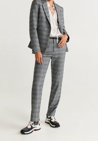 Costume gris à carreaux avec un blazer croisé unique et un pantalon sur mesure, accompagné d'un chemisier blanc et de baskets de sport noires.